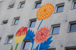 Detailaufnahme des Vikunia Murals in der Mudrastraße für bbd mit großflächigen Blumen und blauen Blättern neben Fenstern.