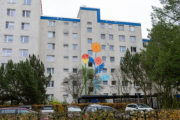 Gesamtansicht der Wohnfassade in der Mudrastraße mit Vikunia Mural für bbd, florales Motiv als farbiger Akzent auf grauer Fassade.