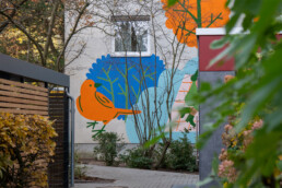 Blick durch Sträucher auf das Vikunia Mural in der Mudrastraße für bbd mit orange blauem Vogelmotiv auf einer Hofwand.