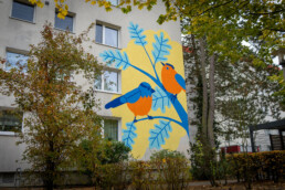 Vikunia Mural in der Mudrastraße für bbd mit zwei stilisierten Vögeln auf gelber Fläche an einer Wohnfassade, umgeben von Bäumen.