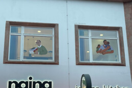 Fensterillustrationen von Vikunia am Dokumentationszentrum mit gezeichneten Figuren in den Innenräumen.
