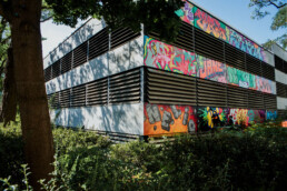 Eckansicht des Parkhauses in Berlin Staaken mit dem großflächigen Mural von Lukas Zeilinger zwischen Bäumen und Grünflächen