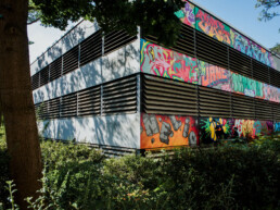 Eckansicht des Parkhauses in Berlin Staaken mit dem großflächigen Mural von Lukas Zeilinger zwischen Bäumen und Grünflächen